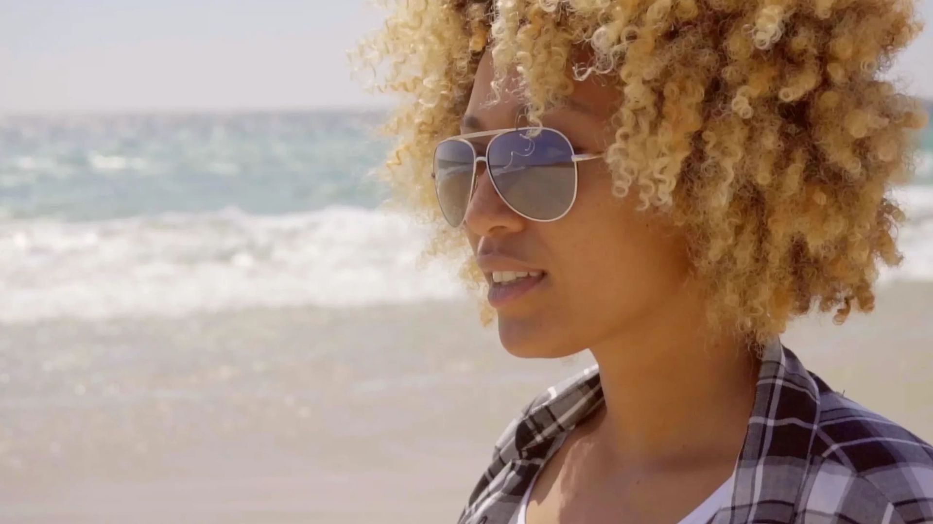 Eine Person mit lockigem blondem Haar trägt eine Sonnenbrille und blickt an einem sonnigen Strandtag auf die Meereswellen.