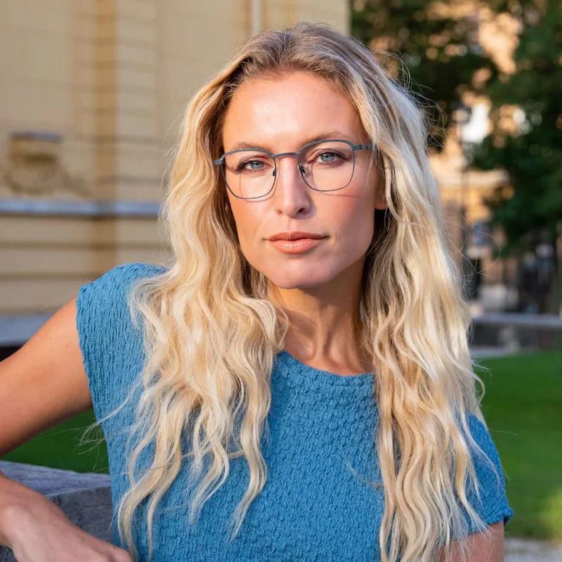 Eine Person mit langen, welligen blonden Haaren, die ein strukturiertes blaues Oberteil trägt, posiert im Freien vor dem Hintergrund von Grünpflanzen und einem historischen Gebäude.