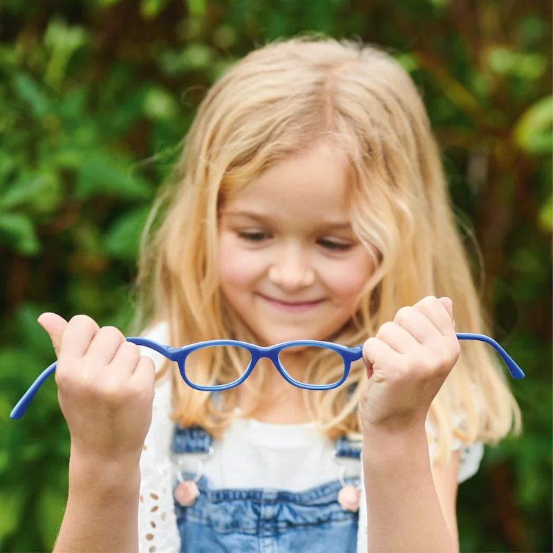 Ein Kind mit blonden Haaren hält eine hellblaue Brille in der Hand und steht vor einem üppig grünen Hintergrund.
