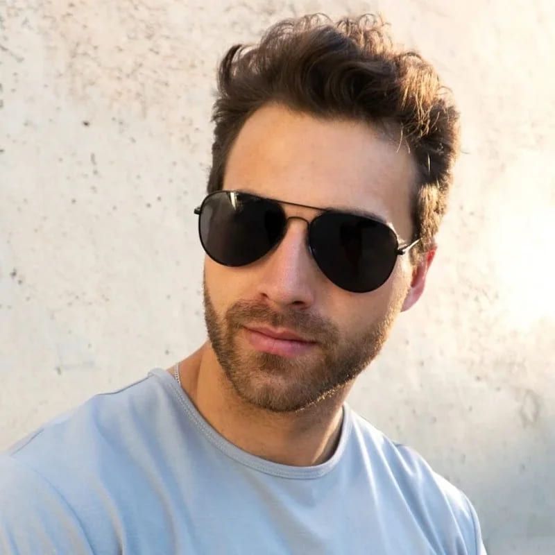 Ein junger Erwachsener mit lockigem Haar und einem hellblauen Hemd posiert vor einem strukturierten weißen Hintergrund und strahlt dabei eine lässige Atmosphäre aus.